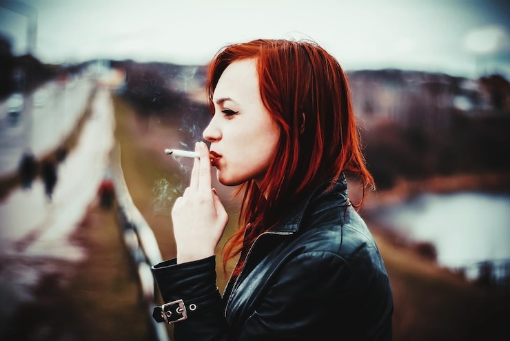 A woman with red hair smoking a cigarette outdoors